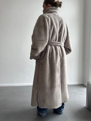 Kvinne iført en myk, beige MEOTINE kåpe med belte, sett bakfra i et lyst rom med minimalistisk interiør.