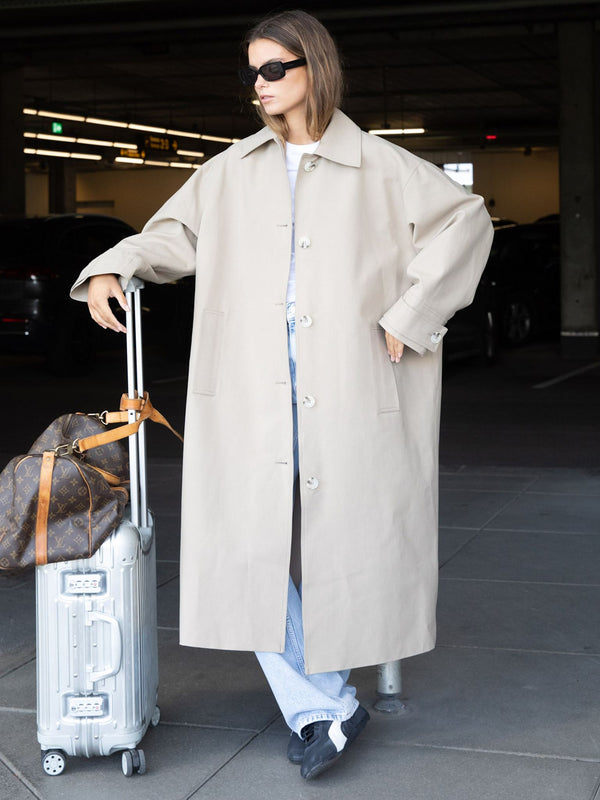 Dame i stilig beige frakk fra MEOTINE står ved en koffert i parkeringshus. Elegant og moderne stil.