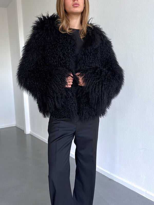 Kvinne iført en svart, fluffy jakke fra MEOTINE, kombinert med svarte bukser, står i et lyst rom med hvite vegger.