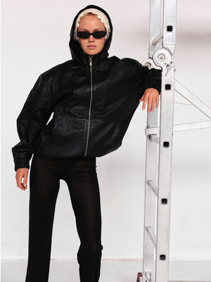 Modell iført svart MEOTINE-jakke, med solbriller, står ved en stige i studio.